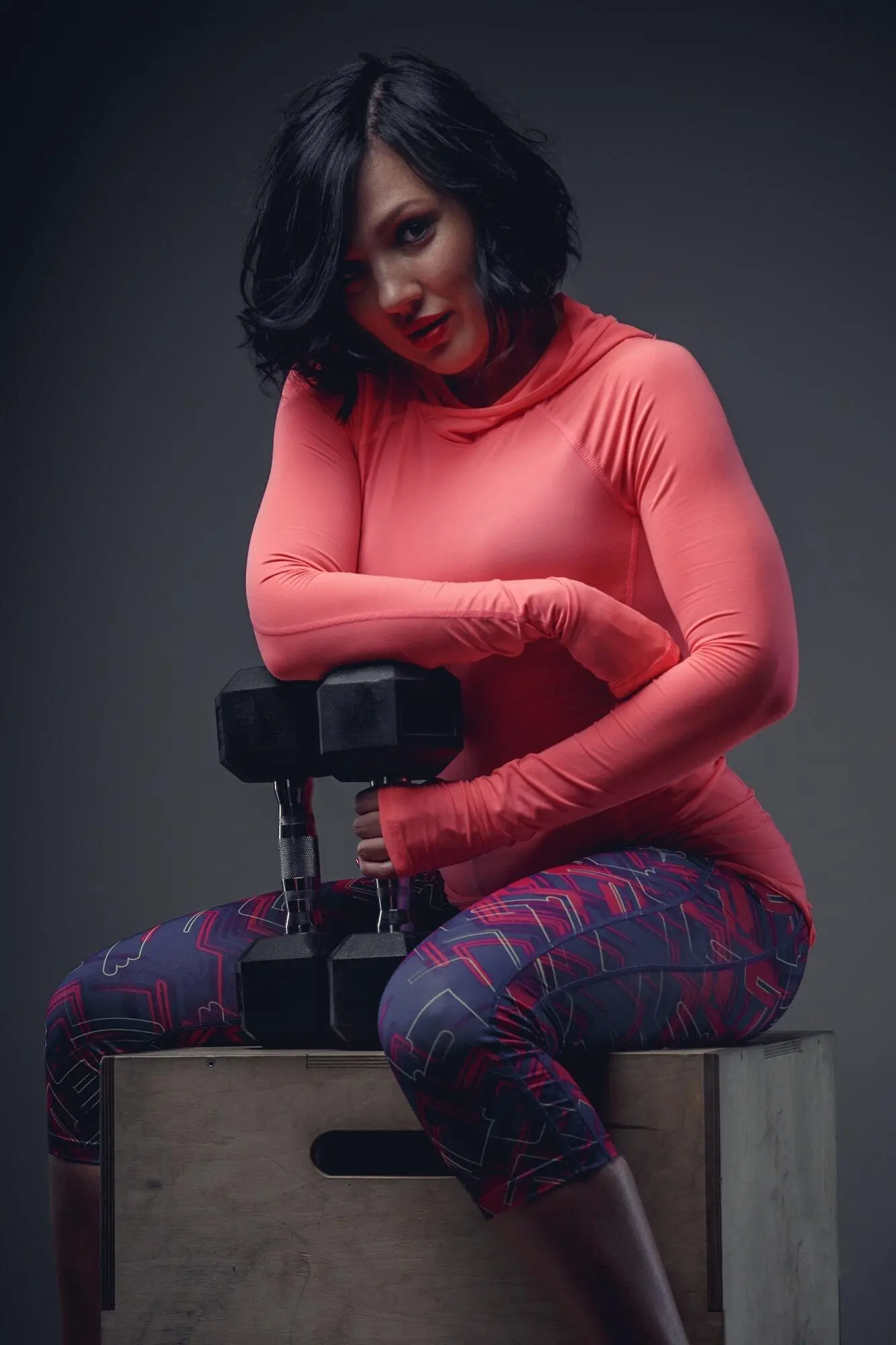 Sportliche brünette Frau in einem rosa T-Shirt sitzt auf einer Holzkiste und hält eine Hantel.