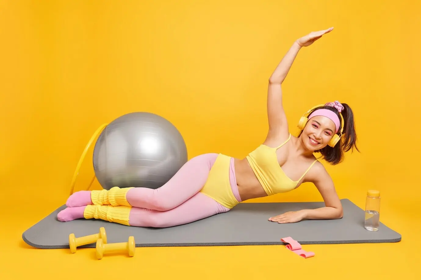 Eine Frau mit östlichem Erscheinungsbild liegt auf einer Fitnessmatte, macht einen seitlichen Plank, hält den Arm nach oben gestreckt, hört Musik über Kopfhörer, trägt Sportbekleidung, hält sich fit und benutzt Sportgeräte.